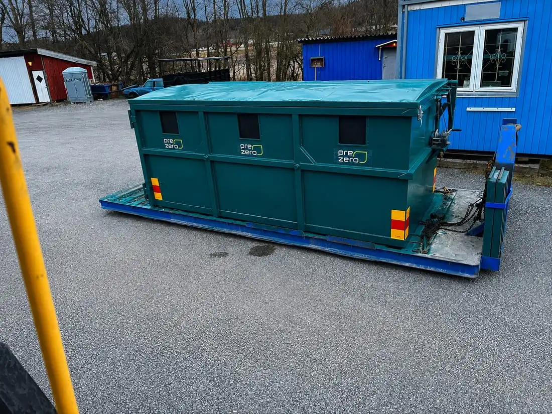 Mörkgrön container med kapell över och tre inkastluckor synliga på långsidan, uppställd på ett blått lastväxlarflak på gården.
