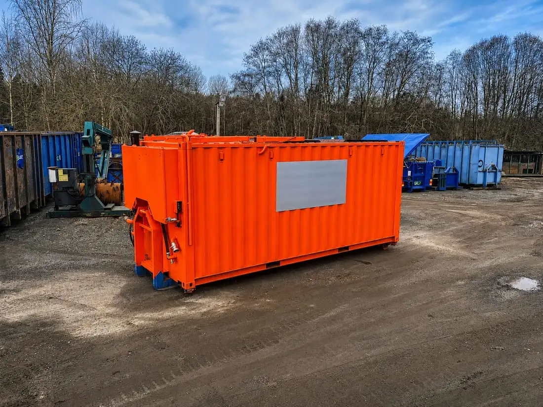 Specialbyggd orange klistertank på lastväxlarram, med hydraulik i fronten, uppställd på gården bland blå containrar.